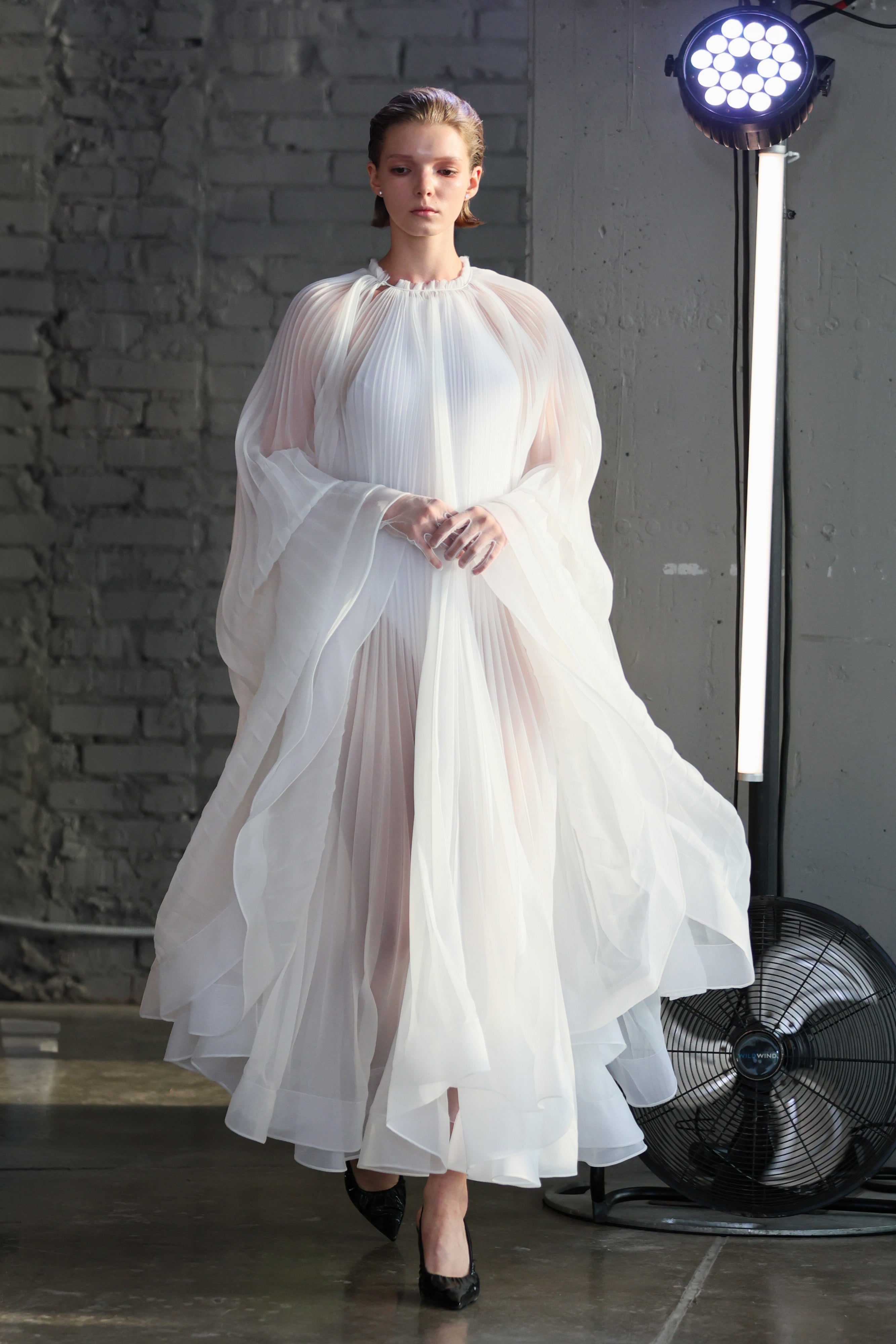Virgin Dress by J'amemme — model wearing white pleated bridal gown backstage with studio lighting