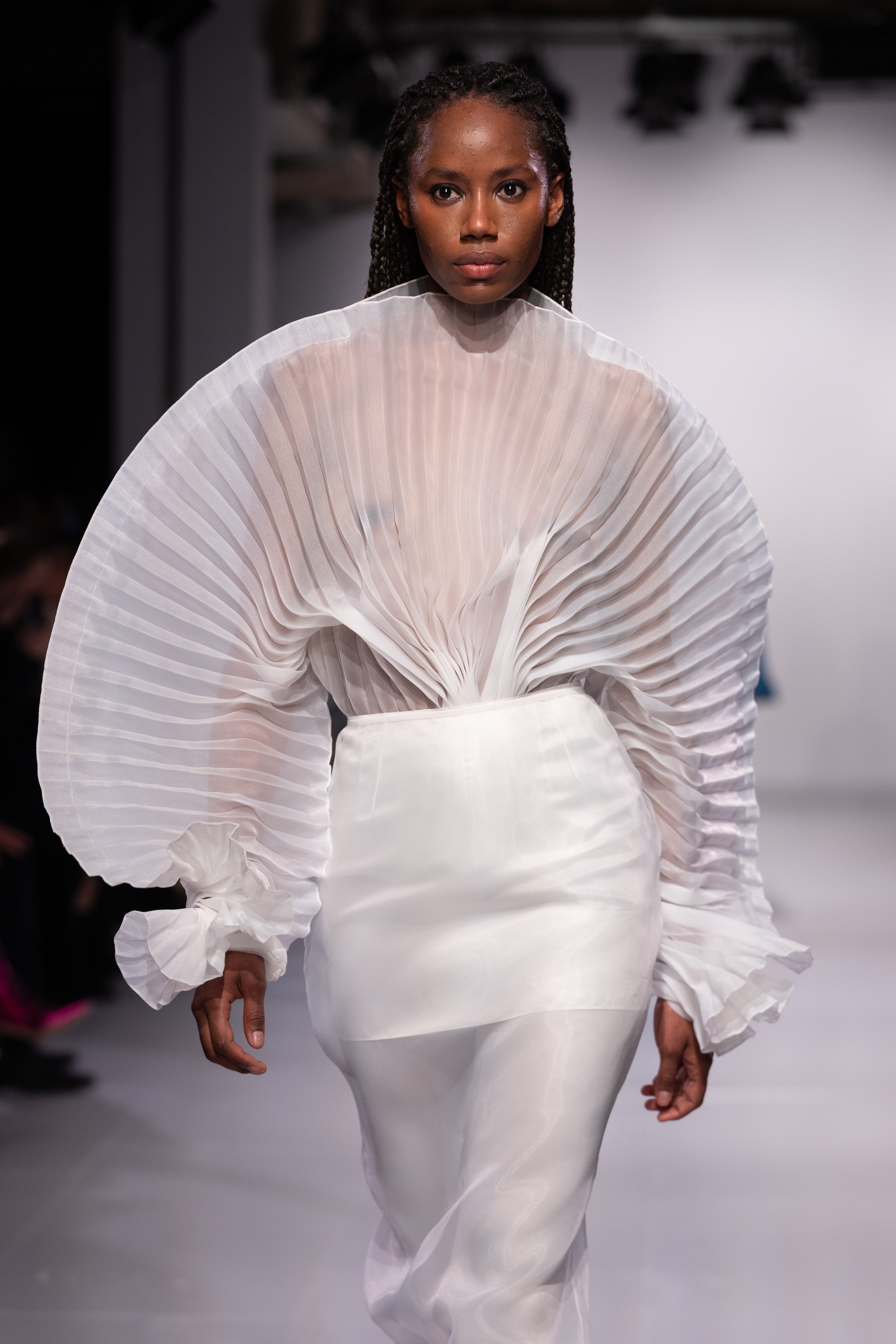 Fleur Set by J'amemme — close-up runway shot of model wearing white pleated bridal top with sculptural sleeves and matching skirt