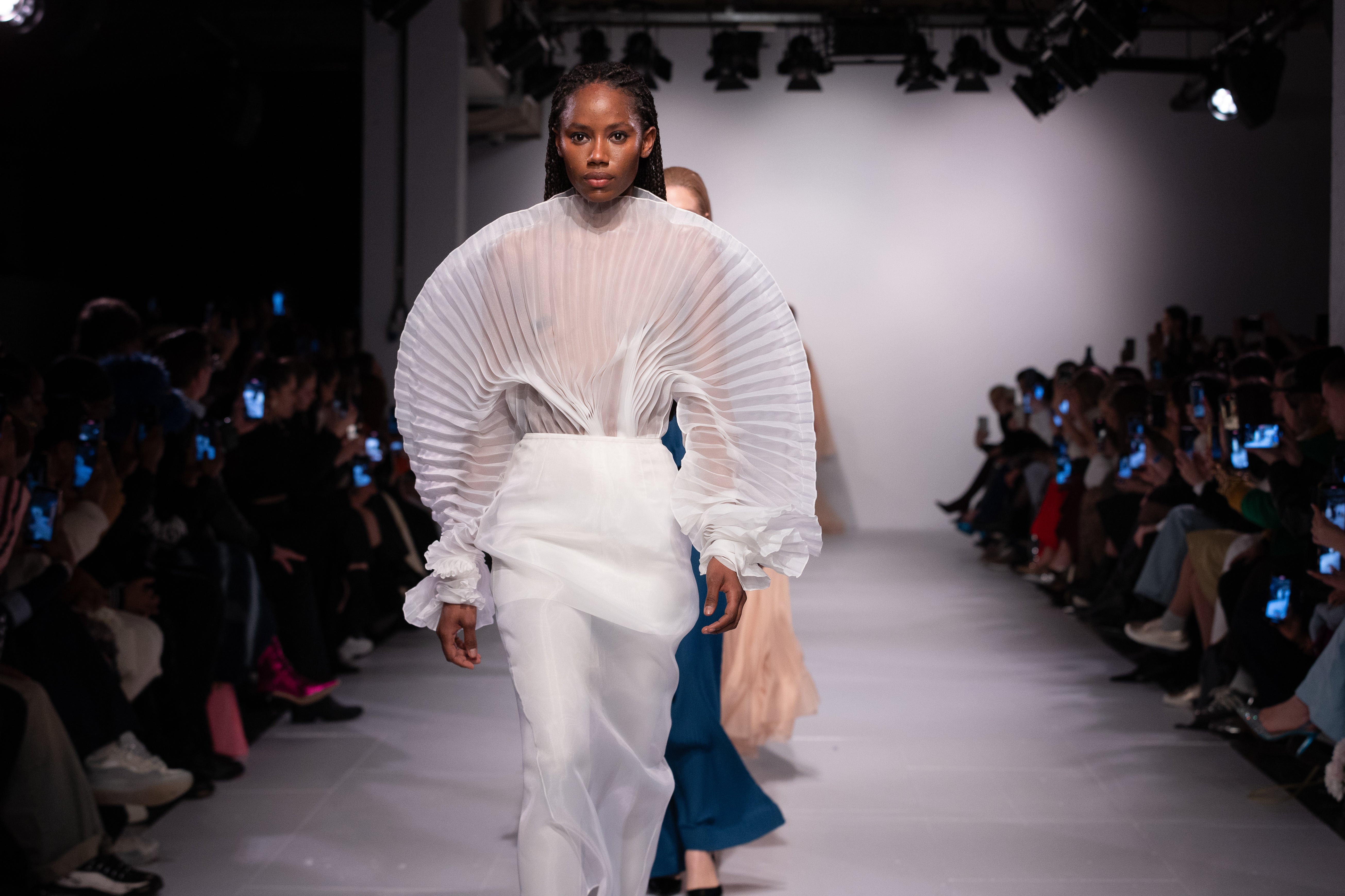 Fleur Set by J'amemme — model walking runway in white pleated bridal two-piece with audience, wide editorial shot